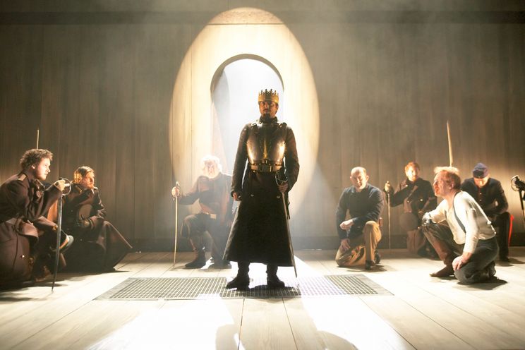Man stands in armour onstage surrounded by kneeling people